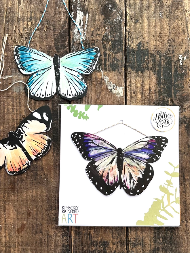 Hand Painted Butterflies a Colourful Linocut Hanging Decoration In multiple colours and black packaging is shown with some leaves on a square card background in an eco friendly compostable cellophane bag to protect it