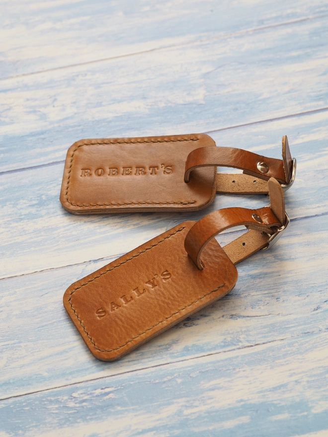 Personalised leather luggage tag. Two tan brown leather luggage tags, each personalised with a name. One is stamped with SALLY'S the other is stamped with ROBERT'S
