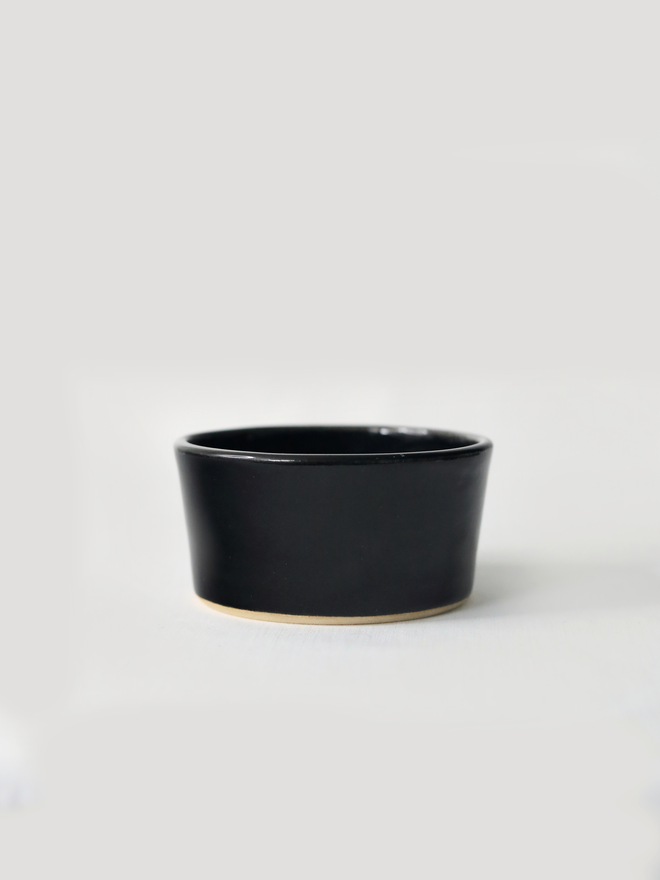 A small handmade ceramic side bowl in glossy jet black glaze, with a tapered sides and a wide open rim, photographed against a plain light grey background