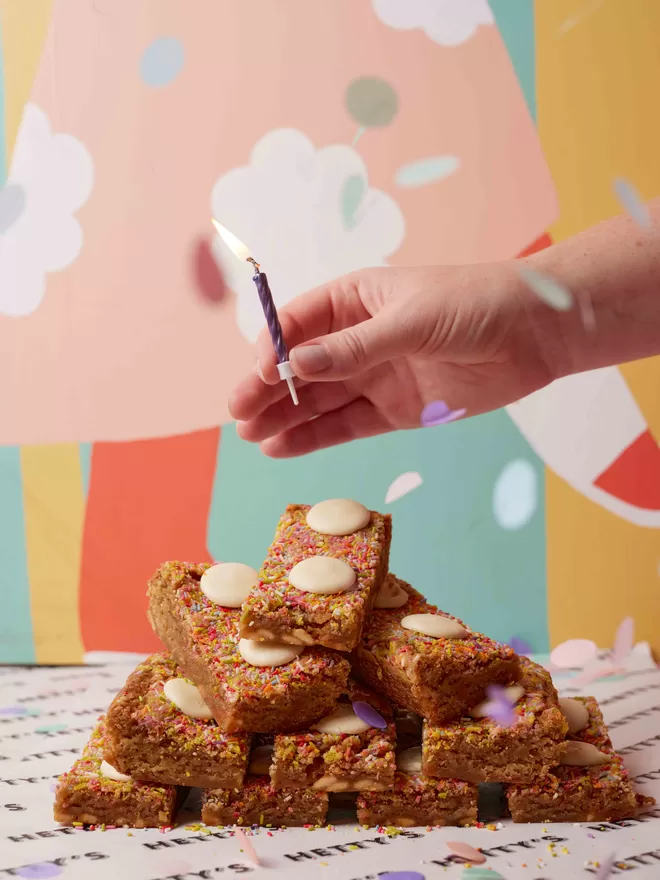 Funfetti blondies with sprinkles and white chocolate buttons stacked for a Birthday celebration with hand holding candle to place on top and confetti in the air