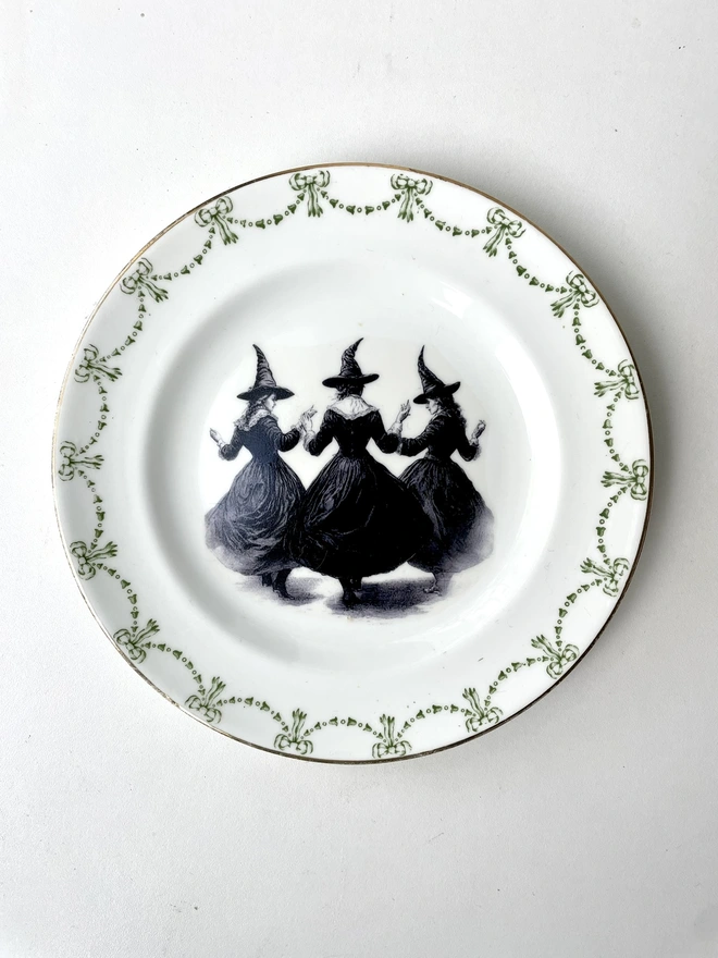 a coven of 3 witches dance hand in hand, the image is victorian in black and white and is printed directly onto an ornate vintage plate