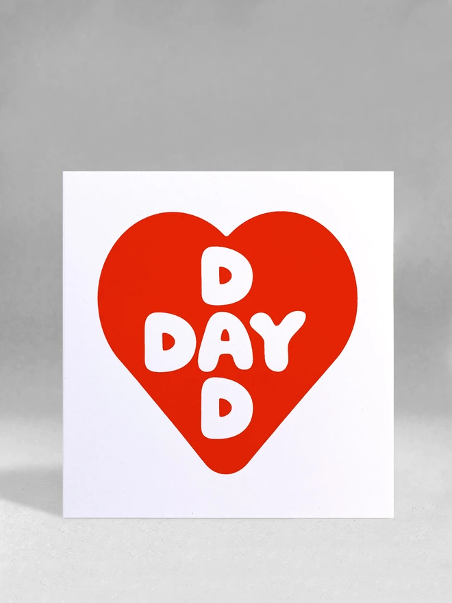 Red screenprinted heart contains intersecting words Dad and Day, a square card stood in a white studio backdrop.