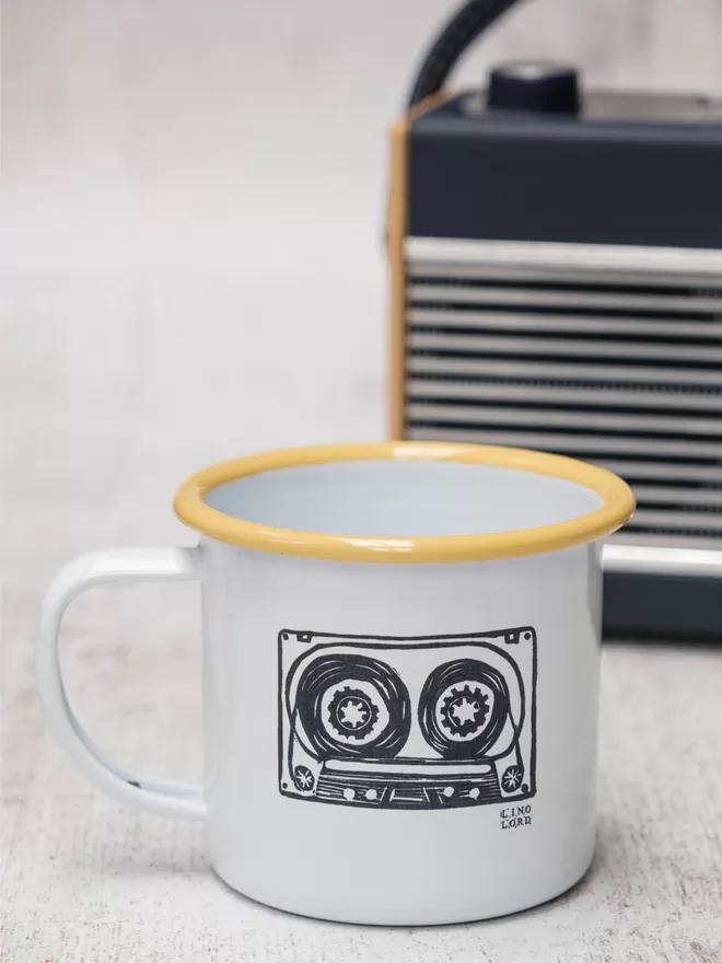 Picture of a White Enamel Mug with a Yellow Rim with a cassette tape design etched onto it, taken from an original Lino Print