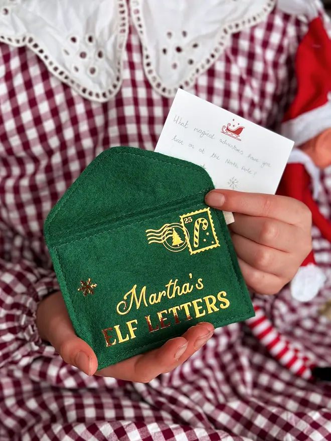 A child wearing a red gingham dress is holding a handmade green felt envelope with golden words reading "Martha's elf letters" and a candy cane design stamp in the corner.