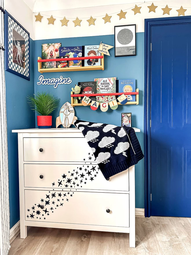 A white chest of drawers with black star decals is situated in a modern space themed nursery painted a vibrant blue. Star decals are stuck on to the front of the chest of drawers cascading down the front. A black and white storm cloud blanket is folded on top on the top.There are bookshelves with childrens books above the drawers and a space rocket sits on the top alongside a wall decal that reads ‘Imagine’.