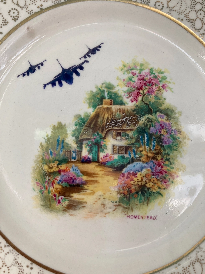 A gold and white china plate featuring a cottage wClose up of A gold and white china plate featuring a cottage with fighter jets in the sky.