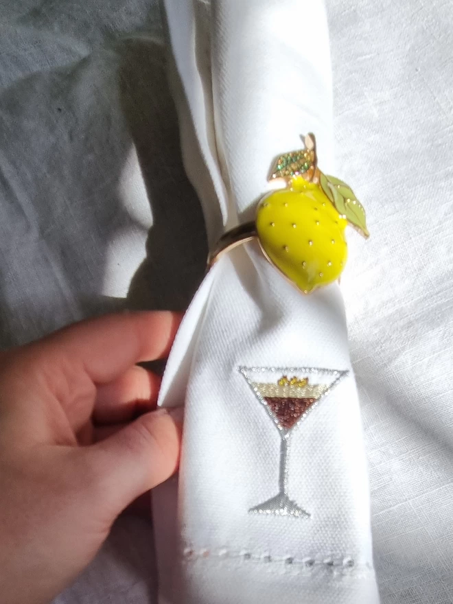 a white napkin with an espresso martini embroidered on it. it is rolled up in a napkin ring