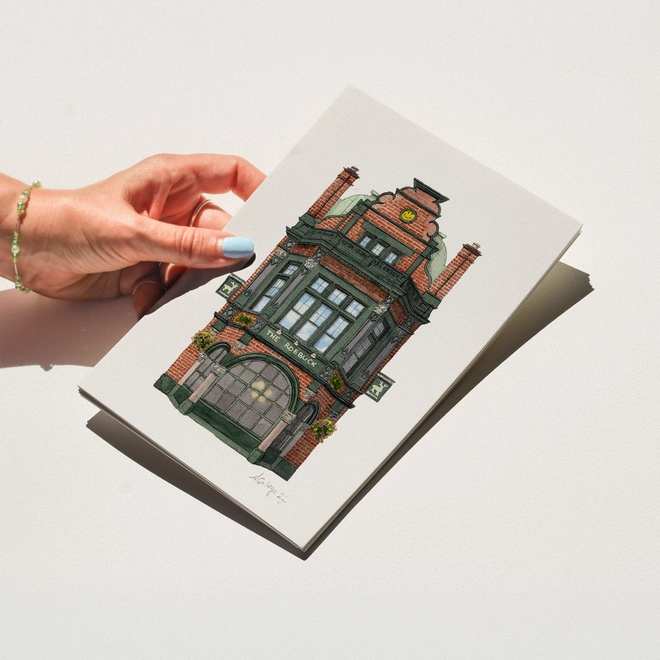 A hand with blue nail polish holding a print featuring an illustration of The Roebuck pub against a white background.