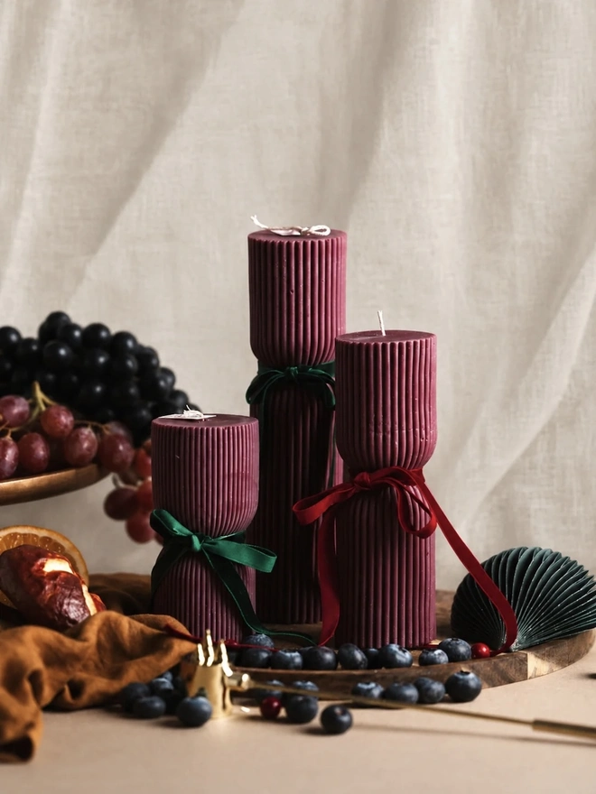 Burgundy Christmas Cracker Pillar Candles (Set Of 3). Photo 1: Three ribbed burgundy pillar candles in graduating heights, each with a cinched waist tied with a green or red velvet bow, on a wooden board with blueberries, grapes and a paper fan ornament. The rich jewel-toned candles make a luxurious centrepiece for Christmas tables.