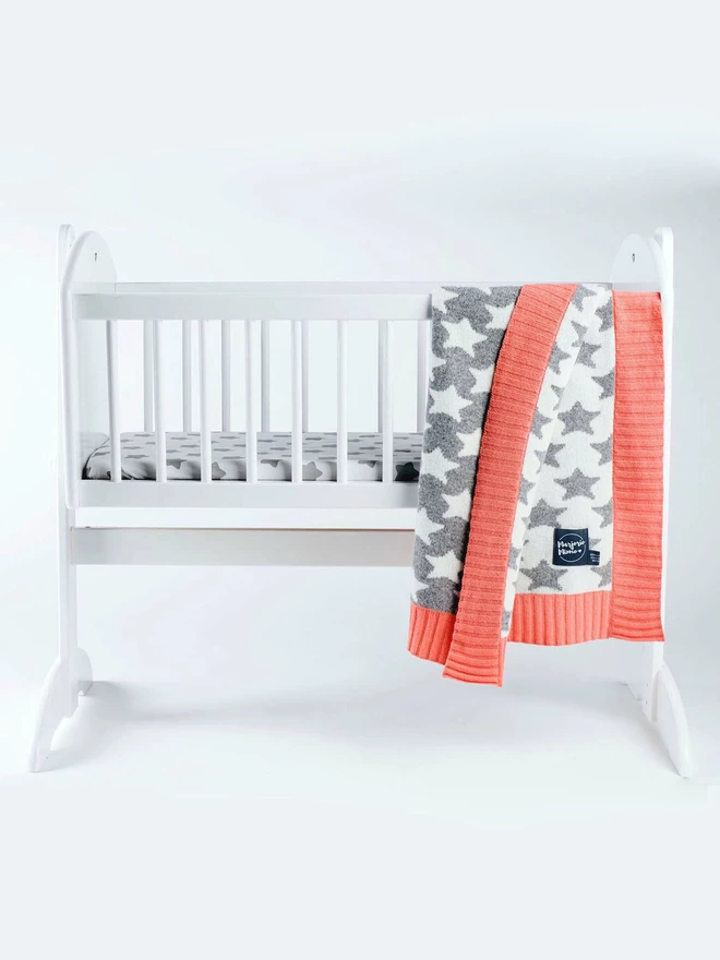 A product shot of a white rocking crib against a white background. A grey and white star pattern baby blanket with coral pink ribbed trim is draped over the side of the crib showing the front and reverse colorway.