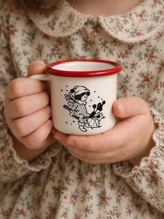 a childs christmas enaml mug being held in a childs hands featuring an illustration of a little fairy pixie sat on a christmas holly lead branch in the snow