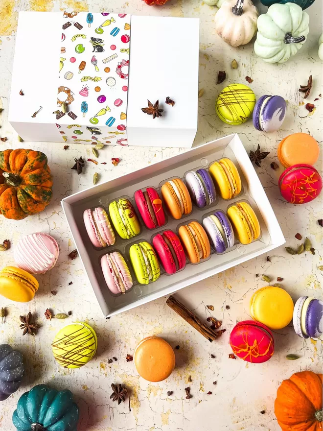 A box of colourful autumn macarons in a white gift box surrounded by colourful pumpkins on a table 