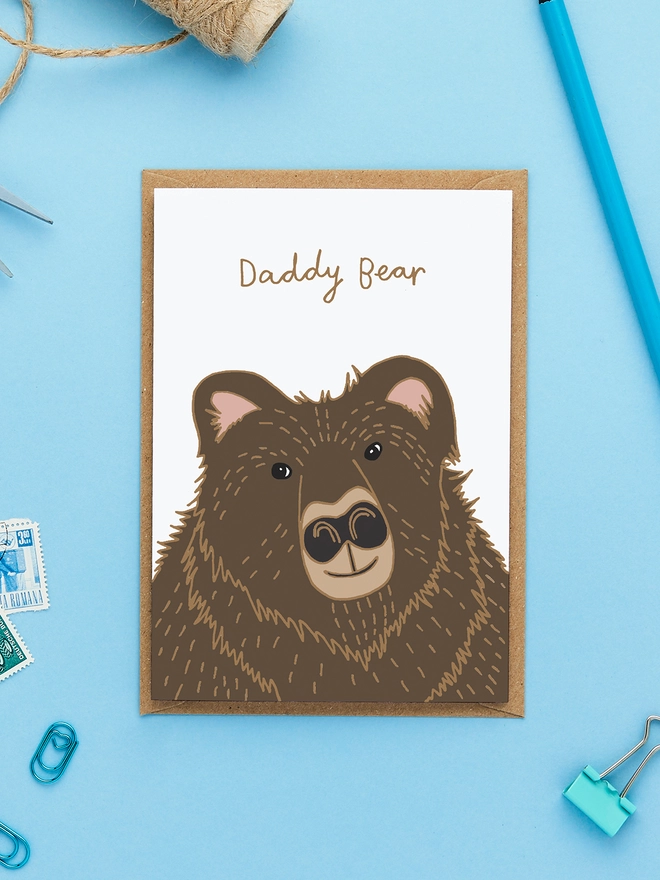 Greeting card featuring an illustrated brown bear with the caption 'Daddy Bear', styled flat lay with blue background, kraft envelope, stamps, and desk accessories.