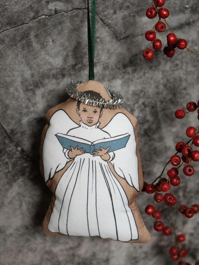 a brown skinned afro caribbean angel christmas tree decoration made from illustrated printed fabric and stuffed with plush hung with ribbon