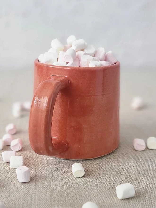 Ceramic Standard Mug, a colourful ceramic mug sitting on a plain surface against a grey backdrop. It is accompanied by marshmallows. 