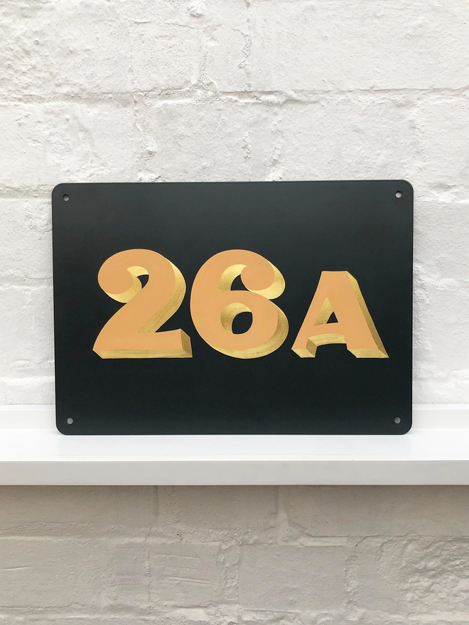 Hand painted house number 26A in pale orange and gold leaf on anthracite grey metal plaque, against a white brick wall. 