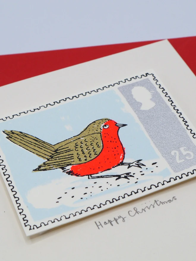 Robin Stamp Christmas Card, a white Christmas card with a colourful bird design in the centre on top of a red envelope on a plain surface. 