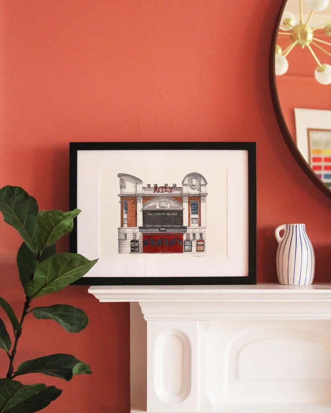 A framed print featuring an illustration of The Ritzy Cinema displayed on a white mantelpiece against a terracotta-colored wall.