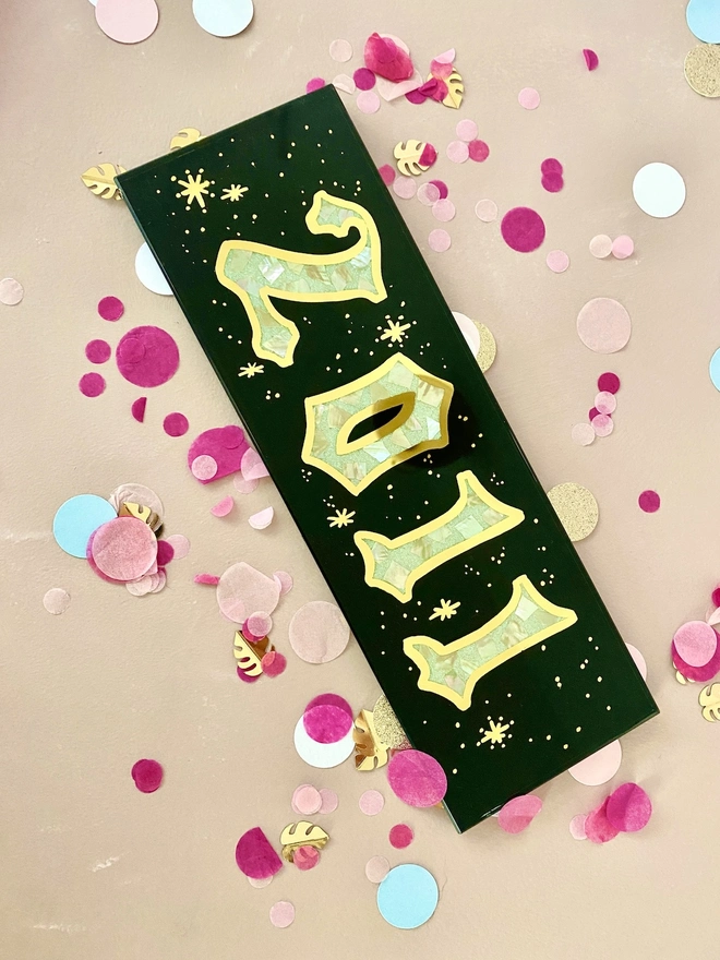 Personalised Year Date Sign, a black and gold personalised year sign sitting on a plain surface joined by confetti. 