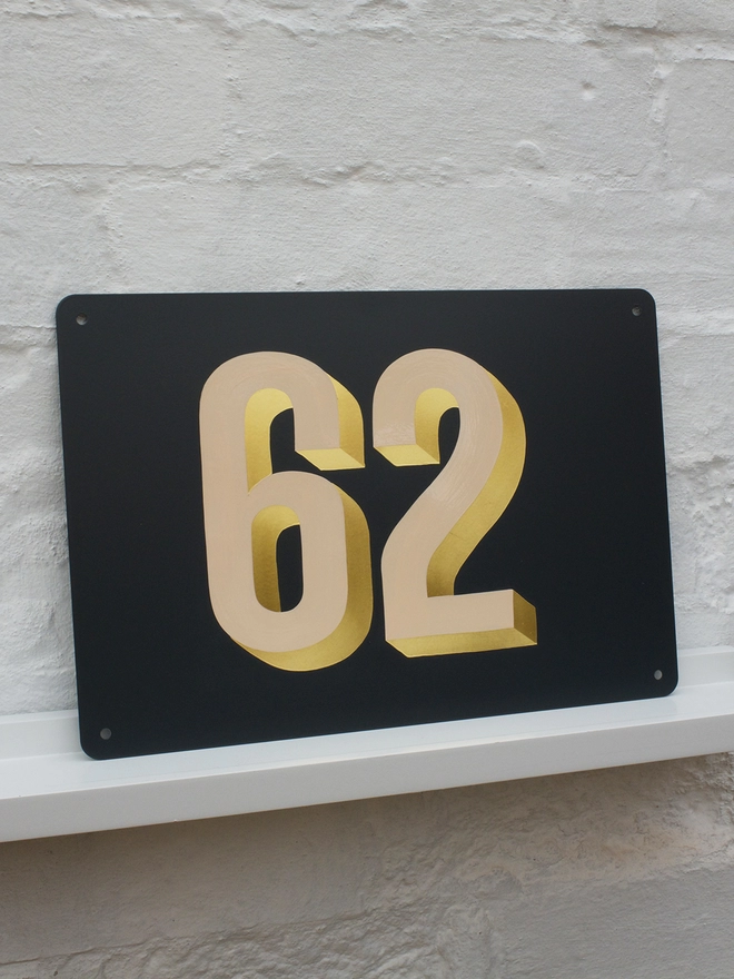 Peach and gold leaf house number 62, on anthracite grey metal plaque, against a white brick wall.