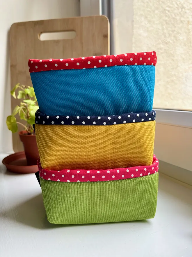 A stack of 3 fabric baskets in green, yellow & blue on a windowsill, all with polka dot linings.