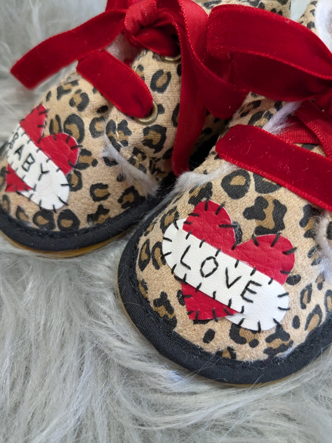 A pair of leopard print baby booties, lined with fur and with red velvet ribbon laces. A red heart with a white scroll adorns each booty. One says BABY, the other, LOVE in black stitches