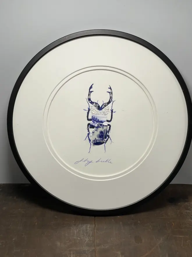 Hand-drawn, painted, mosaic clay, blue Willow Tree pottery insect on white double mounted card, in a black circular frame.