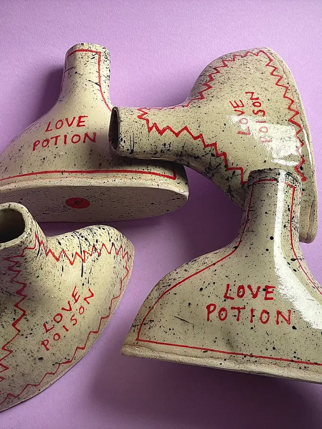 Love Potion/ Poison Bottle Vase, a group of ceramic vases that each say 'love poison/ love potion' grouped together on a purple background. Each is stamped with a signature red dot on the bottom. 