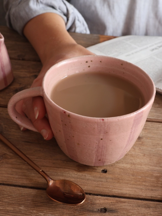 A female hand holding the handmade Ceramic Stoneware Mug in Pink Stripe. The mug sits on a wooden surface next to a newspaper and a brass spoon.  