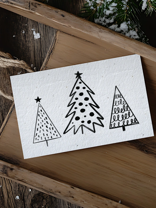 A Christmas card with three black and white doodle trees placed on a wooden sleigh with wintery greenery.