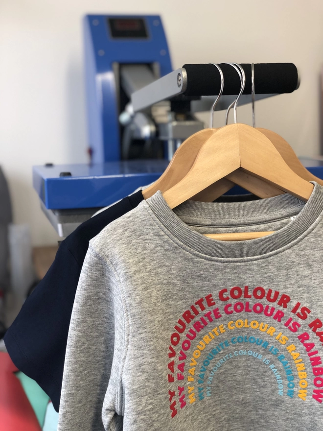 a 'Rainbow' Sweatshirt by The Faraway Gang with a rainbow on it displayed on a hanger..