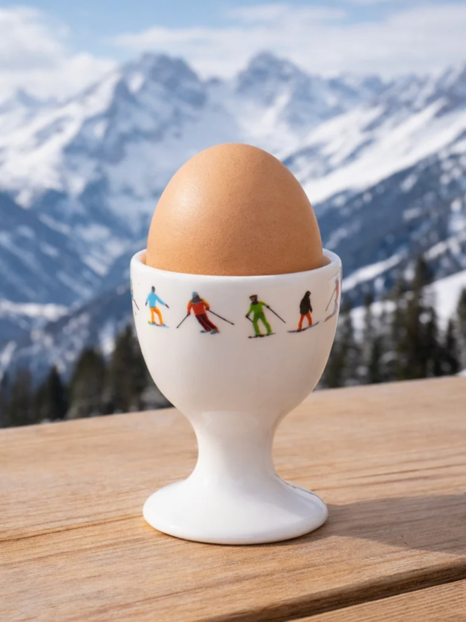 Ski Chain Egg Cup. A white egg cup patterned with a chain of multicoloured skiers on its rim. It is set against a mountain backdrop and holds a brown egg.