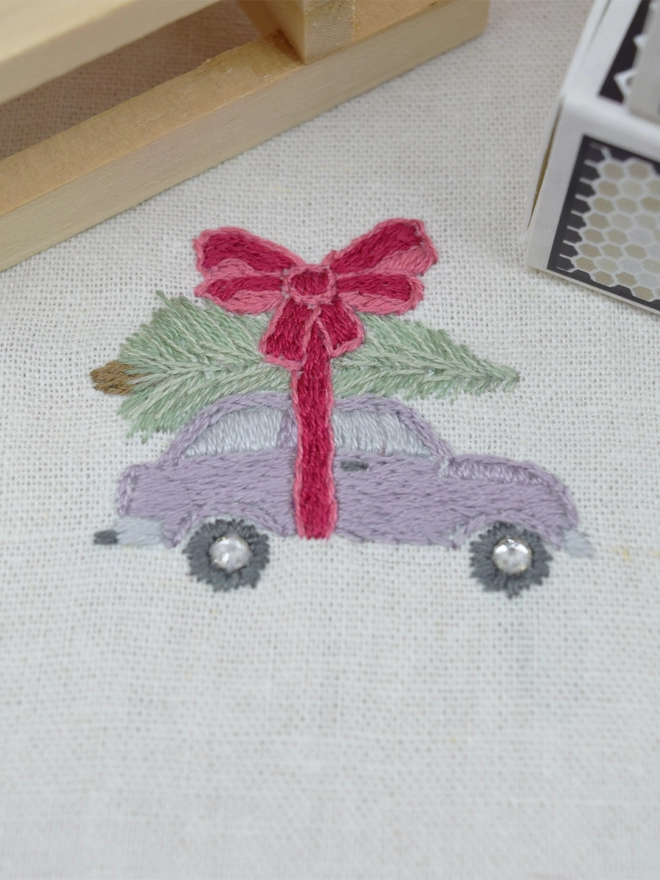 An embroidered lilac purple car carrying a Christmas fir tree on the roof, strapped in with a giant pink bow.