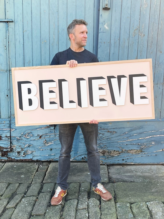 A person holding a painted wooden sign reading BELIEVE on a pale pink background, standing in front of a blue door with cracked painted