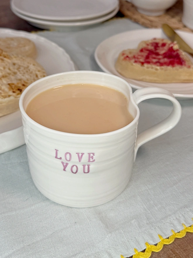 hand thrown porcelain cream mug with pink lustre text on the side that says LOVE YOU. The mug is sat upon a sage table cloth amongst a tea time scene of heart shaped crumpets