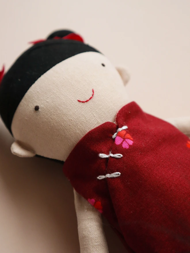 A handmade doll with black hair and red bows, wearing a red dress with floral accents, sits on a green box. Miniature wooden tea sets and kitchenware are arranged nearby, including a small table, cups, and a teapot.