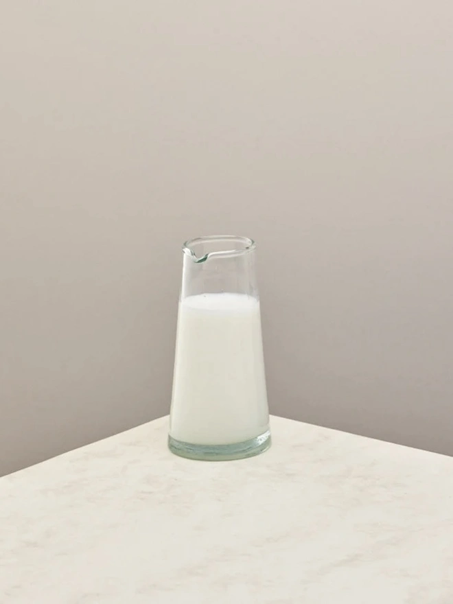 Tall Jug, a tall glass jug sitting on a plain counter top against a white backdrop. It is filled by milk.