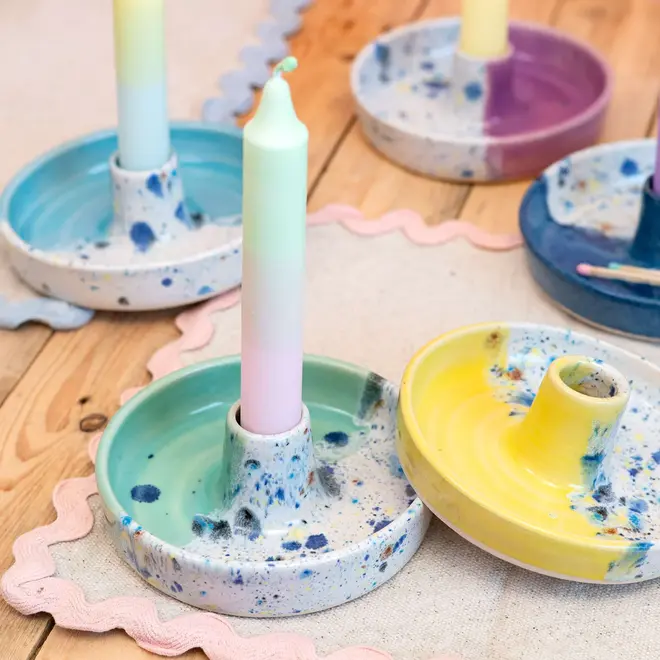 Shoreline Candle Stick Holder, a collection of colourful candle stick holders sitting on a wooden surface against a pink backdrop. They are accompanied by colourful napkins. 