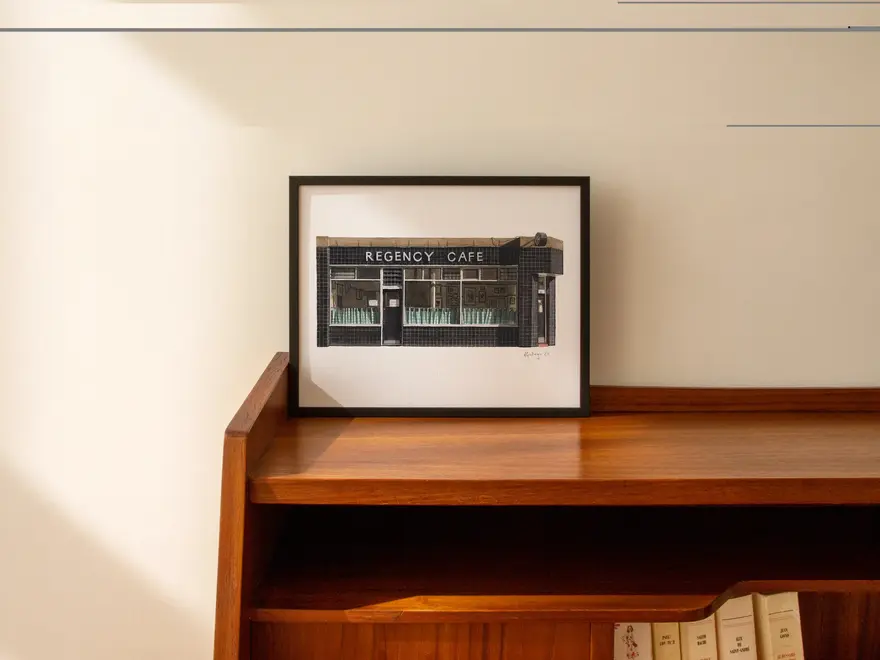 Pimlico Regency Cafe Giclée Print (Unframed) Victoria, a print of a cafe in a black frame sitting on a wooden surface against a plain backdrop. 