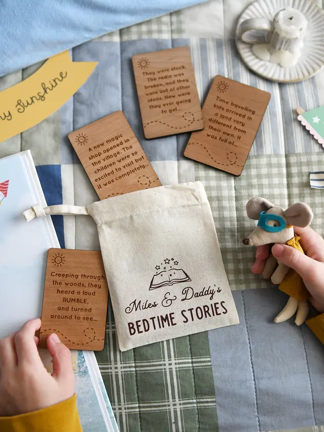 Personalised bedtime story set for dads and children featuring engraved wooden story prompt cards and a drawstring cotton bag reading 'Miles & Daddy’s Bedtime Stories', displayed on a patchwork quilt with a soft toy, picture book, and a young child holding one of the cards.