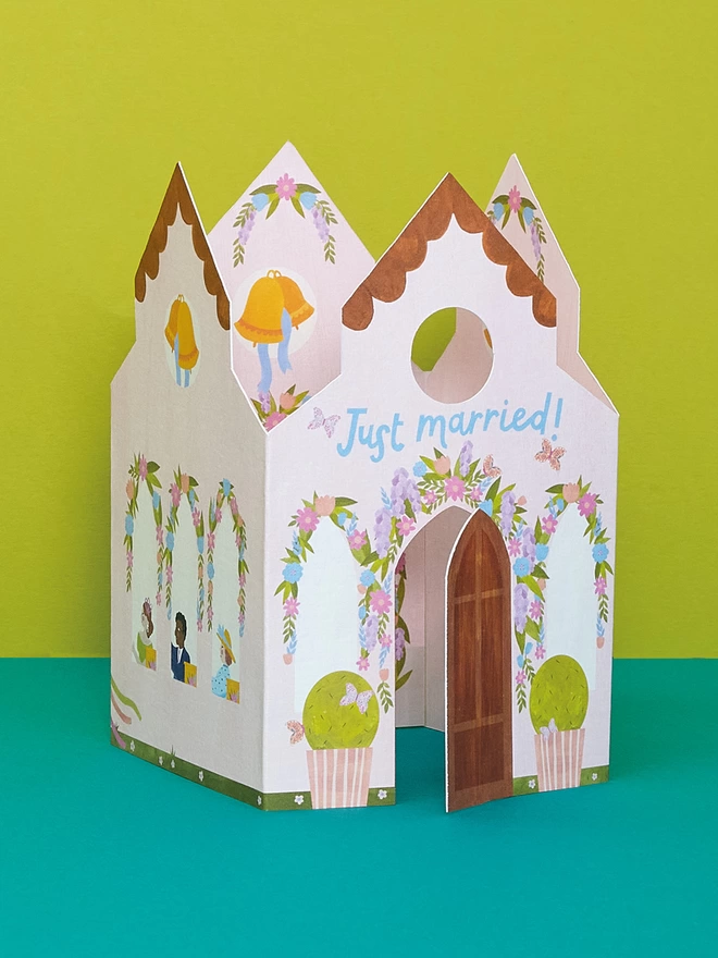 The 3D fold out 'Just Married' church card features die cut door which opens out. The church is covered in painted flowers and through the windows we can see wedding guests ready to celebrate the happy couple