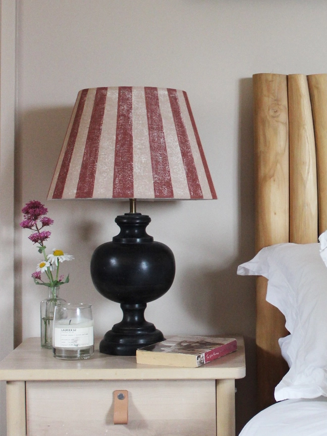 Empire lampshade with bold burgundy and pink stripes, styled on a black table lamp beside a bed with a wooden Tikamoon headboard. A colourful lampshade and classic striped lampshade for bedrooms.
