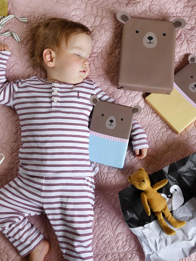 A baby wearing red and white striped pyjamas is asleep on a pink quilt with three gifts wrapped up as teddy bears.