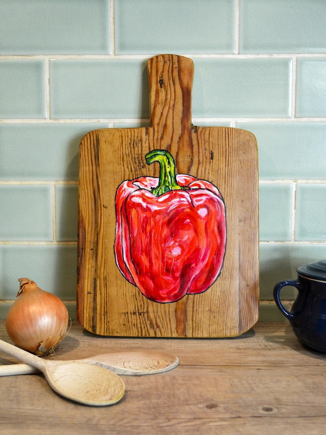 Wooden chopping board with handpainted design of a red pepper standing against a kitchen wall