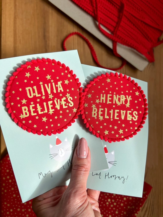 Red christmas 'Believe' badges with gold stars and a red pom pom trim and a child's name 