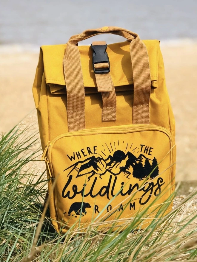 'Wildings' Children’s Recycled Roll Top Back Pack, a colourful backpack sitting on a shore in front of a sea. 