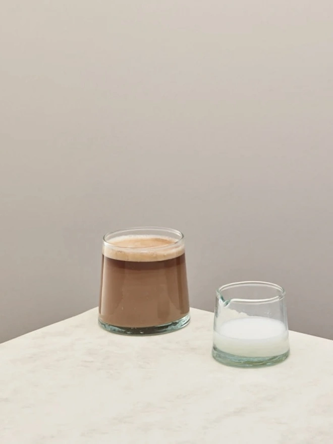Low Jug, a small glass jug sitting on a counter top against a plain backdrop. It is filled by milk and accompanied by another glass. 