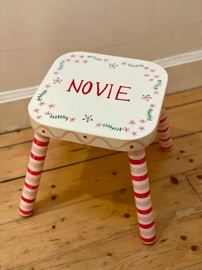 personalised hand-painted nursery stool. a small white stool with red and pink striped legs, painted with 'novie' on top.