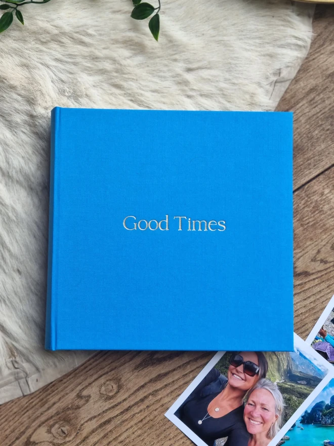 A sky blue photo album printed with the words 'Good Times' rests on a wooden floor and a rug, next to family photos.