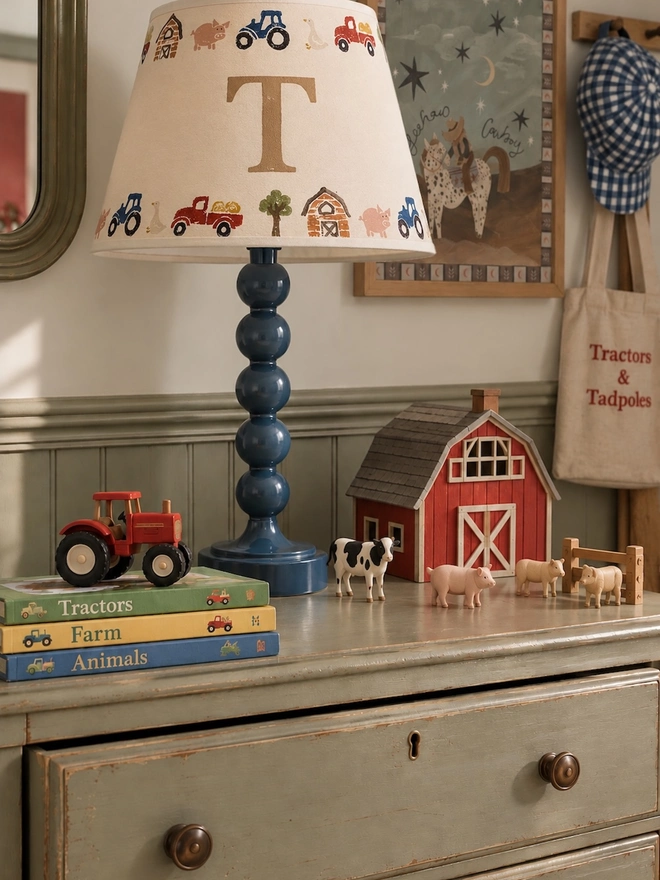 Studio Zef personalised farmyard lampshade with initial T, styled on a glossy navy blue bobbin table lamp in a charming countryside-inspired children's bedroom. The cream hand-printed lampshade features colourful tractors, pigs, geese and barns, displayed alongside a miniature red wooden barn, toy farm animals, tractor books and vintage-style artwork on a sage green chest of drawers.